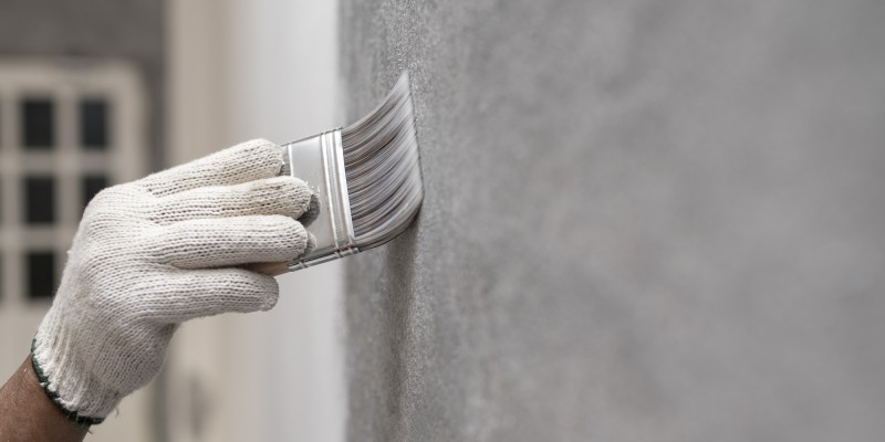Hand mit weißem Arbeitshandschuh streicht mit einem Pinsel graue Wand.