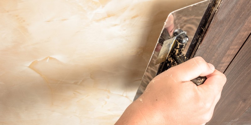 Hand trägt mit Glättkelle eine marmorähnliche Spachteltechnik auf eine Wand auf.